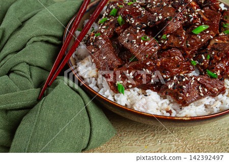 crispy beef, with green onions and sesame seeds, on a rice pad, in a bowl with chopsticks, on a textured surface, no people crispy beef, with green onions and sesame seeds, on a rice pad, in a bowl with chopsticks, on a textured surface, no people 142392497