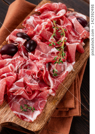Freshly cut dried meat, served with olives and herbs, on a wooden chopping board, no people, Freshly cut dried meat, served with olives and herbs, on a wooden chopping board, no people, 142392498