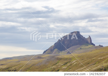 Icelandic mountain stands tall under a cloudy sky with green hills around it in a remote area Icelandic mountain stands tall under a cloudy sky with green hills around it in a remote area 142392837