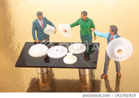 Miniature people discuss gears on a table in an industrial setting during daytime Miniature people discuss gears on a table in an industrial setting during daytime 142392919