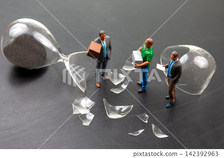 Miniature figures gather around broken hourglass with scattered pieces on black surface Miniature figures gather around broken hourglass with scattered pieces on black surface 142392963