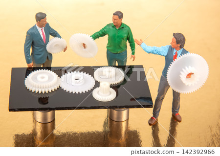 Miniature figures interacting with gears on a table in a creative workspace setting Miniature figures interacting with gears on a table in a creative workspace setting 142392966