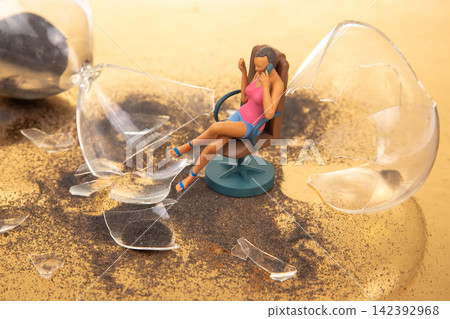 Miniature figure sitting on chair amid broken glass on a table Miniature figure sitting on chair amid broken glass on a table 142392968