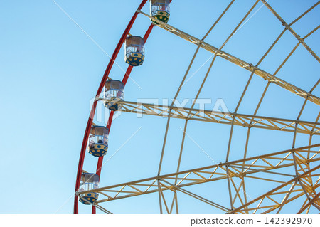 Ferris wheel with cabins against blue sky in an amusement park during sunny day Ferris wheel with cabins against blue sky in an amusement park during sunny day 142392970