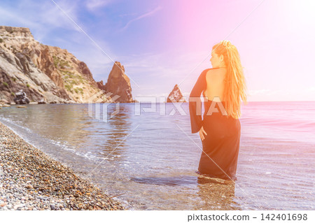 Woman beach ocean standing in shallow water on a pebble beach with majestic coastal cliffs and a sun flare effect 142401698