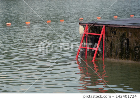 A moody and atmospheric view of a dark lake with safety buoys and a red metal stairs. Concept of a water accident, emergency incident, risk, and rescue operations without people. A moody and atmospheric view of a dark lake with safety buoys and a red metal stairs. Concept of a water accident, emergency incident, risk, and rescue operations without people. 142401724