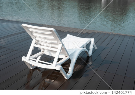 Single plastic lounge chair on a wooden plank surface reflecting calm clear water. Concept of silence, weekend getaway, mindfulness travel, and calm morning atmosphere during summertime. Single plastic lounge chair on a wooden plank surface reflecting calm clear water. Concept of silence, weekend getaway, mindfulness travel, and calm morning atmosphere during summertime. 142401774
