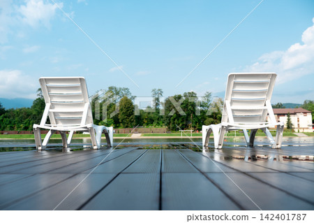 Two white lounge chairs standing on a wet deck with reflections under a blue sky. Peaceful morning view of a poolside terrace, perfect for luxury summer vacation and relaxation concepts. Two white lounge chairs standing on a wet deck with reflections under a blue sky. Peaceful morning view of a poolside terrace, perfect for luxury summer vacation and relaxation concepts. 142401787