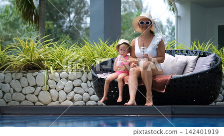 Mother in white swimsuit and straw hat with little daughter in pink swimsuit sitting comfortably in pool lounge chairs drinking fruit cocktails at a resort. Relaxed family concept 142401919