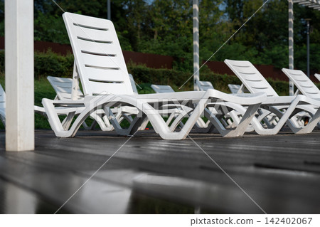 Low angle view of multiple lounge chairs standing on a dark plank deck with reflections. Luxury poolside recreation scene representing peace, summer vacation, and premium relaxation concept Low angle view of multiple lounge chairs standing on a dark plank deck with reflections. Luxury poolside recreation scene representing peace, summer vacation, and premium relaxation concept 142402067