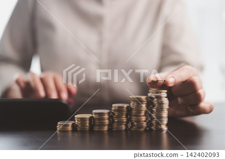 Person adding a coin to rising stacks beside a calculator, symbolizing savings growth, investment planning, budgeting discipline, financial progress, wealth building, and long-term security. Person adding a coin to rising stacks beside a calculator, symbolizing savings growth, investment planning, budgeting discipline, financial progress, wealth building, and long-term security. 142402093