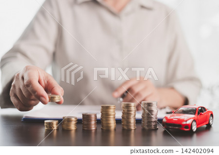 Person adding coins to rising stacks beside a red model car, representing vehicle savings, auto loan planning, down payment growth, budgeting discipline, purchase affordability, and ownership goals. Person adding coins to rising stacks beside a red model car, representing vehicle savings, auto loan planning, down payment growth, budgeting discipline, purchase affordability, and ownership goals. 142402094