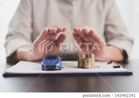 Hands sheltering a blue model car and stacked coins on financial documents symbolize vehicle insurance, asset protection, risk management, ownership security, budgeting, and responsible planning. Hands sheltering a blue model car and stacked coins on financial documents symbolize vehicle insurance, asset protection, risk management, ownership security, budgeting, and responsible planning. 142402241