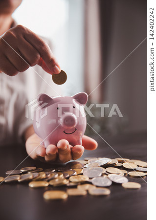 Person holding a pink piggy bank while adding a coin above scattered money, symbolizing hands-on saving, personal finance, household budgeting, financial goals, and future stability. 142402242