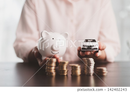 Person holding a white piggy bank and model car above coin stacks, representing vehicle savings, purchase planning, budgeting, investment growth, financial discipline, and future ownership. Person holding a white piggy bank and model car above coin stacks, representing vehicle savings, purchase planning, budgeting, investment growth, financial discipline, and future ownership. 142402243