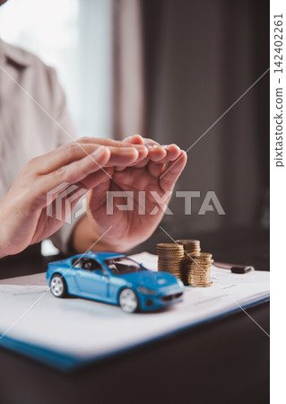 Protective hands surround a blue car model and stacked coins on financial documents, expressing vehicle insurance, asset security, risk management, savings protection, and responsible ownership. Protective hands surround a blue car model and stacked coins on financial documents, expressing vehicle insurance, asset security, risk management, savings protection, and responsible ownership. 142402261
