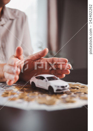 Hands protect a white car model placed on coins and banknotes, representing automobile insurance, financial safety, asset protection, risk control, secure ownership, and emergency planning. Hands protect a white car model placed on coins and banknotes, representing automobile insurance, financial safety, asset protection, risk control, secure ownership, and emergency planning. 142402262