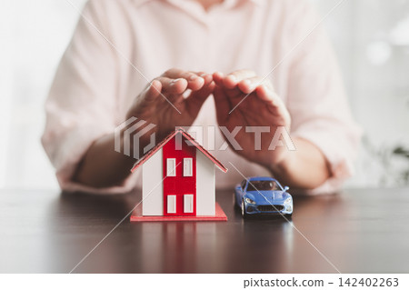 Hands sheltering a red and white model house with a blue toy car, symbolizing property and vehicle insurance, family protection, asset security, risk management, ownership, and customer trust. Hands sheltering a red and white model house with a blue toy car, symbolizing property and vehicle insurance, family protection, asset security, risk management, ownership, and customer trust. 142402263