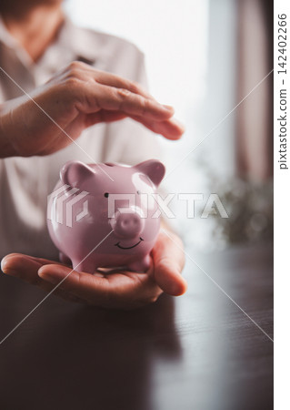 Hands gently protecting a pink piggy bank on a dark table, representing financial safety, savings protection, risk management, insurance, emergency funds, and secure future planning. Hands gently protecting a pink piggy bank on a dark table, representing financial safety, savings protection, risk management, insurance, emergency funds, and secure future planning. 142402266