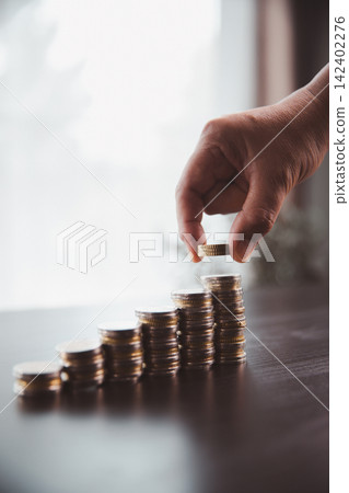 Hand placing a coin on rising stacks of money, symbolizing savings growth, financial progress, investment strategy, wealth building, budgeting discipline, profit, and long-term financial goals. Hand placing a coin on rising stacks of money, symbolizing savings growth, financial progress, investment strategy, wealth building, budgeting discipline, profit, and long-term financial goals. 142402276