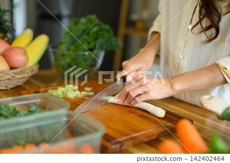 Woman Chopping Green Onion on Wooden Cutting Board with Fresh Vegetables in Home Kitchen 142402464