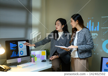 Two Professional Businesswomen Discussing Data Presentation with Charts on Monitor and Projected Dashboard in Office Two Professional Businesswomen Discussing Data Presentation with Charts on Monitor and Projected Dashboard in Office 142402502
