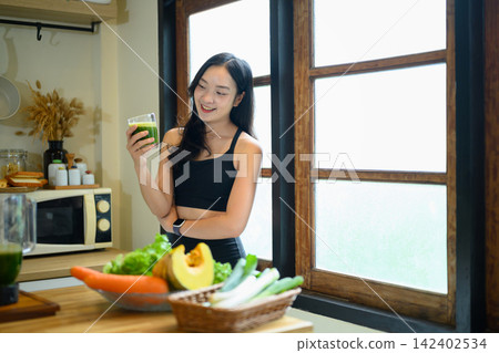 Young Woman in Sportswear Holding Green Kale Smoothie in Kitchen with Fresh Vegetables for Healthy Lifestyle 142402534