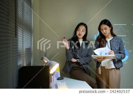 Two Businesswomen Discussing Data Presentation with Charts on Screen and Projector in Modern Office Two Businesswomen Discussing Data Presentation with Charts on Screen and Projector in Modern Office 142402537