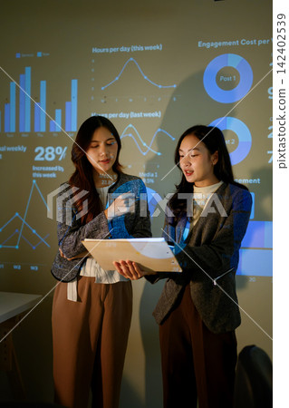 Two Businesswomen Discussing Data Charts During Corporate Presentation with Projected Dashboard Two Businesswomen Discussing Data Charts During Corporate Presentation with Projected Dashboard 142402539