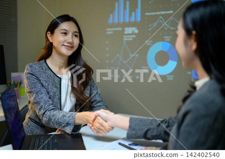 Business Women Shaking Hands After Successful Presentation Meeting with Data Charts Projected in Background Business Women Shaking Hands After Successful Presentation Meeting with Data Charts Projected in Background 142402540