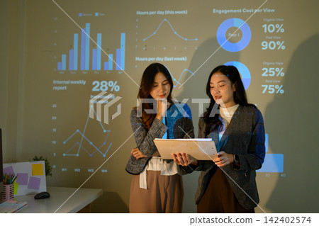 Two Businesswomen Reviewing Data During Office Presentation with Projected Charts and Analytics Dashboard Two Businesswomen Reviewing Data During Office Presentation with Projected Charts and Analytics Dashboard 142402574