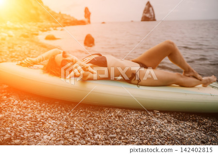 Sunbathing woman, young female relaxing on yellow paddleboard at scenic coastal rocky shore during sunset Sunbathing woman, young female relaxing on yellow paddleboard at scenic coastal rocky shore during sunset 142402815
