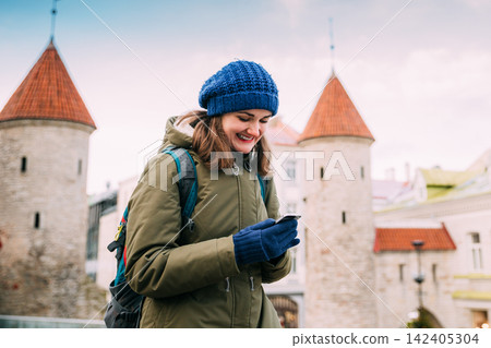 Tallinn, Estonia. Young Caucasian Woman Backpacker Tourist Enjoying Life, Smiling And Using Smartphone Near Landmark Viru Gate. Vacation In Old Town. Girl Surfing Net Internet In Phone 142405304