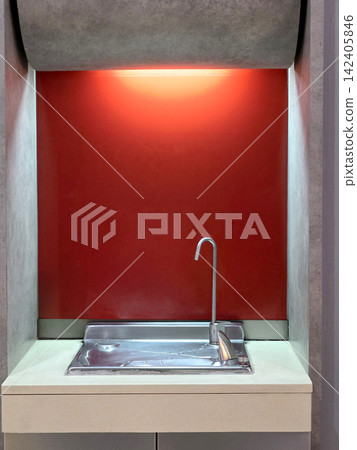 Modern water drinking fountain set in a minimalist wall niche. Features a sleek chrome faucet, stainless steel basin, and a bold red background illuminated by a warm orange glow from the top light. 142405846