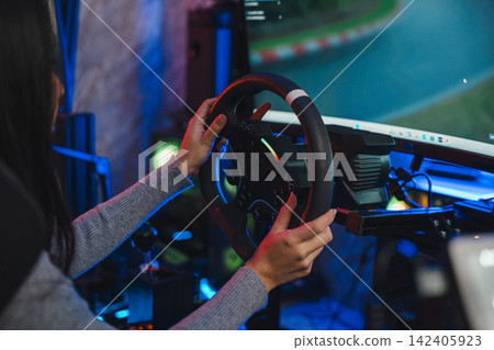 Woman playing game on car simulator while holding steering wheel in dark room with colorful neon lights, enjoying virtual drive during hobby time at home, focused on digital race track screen Woman playing game on car simulator while holding steering wheel in dark room with colorful neon lights, enjoying virtual drive during hobby time at home, focused on digital race track screen 142405923