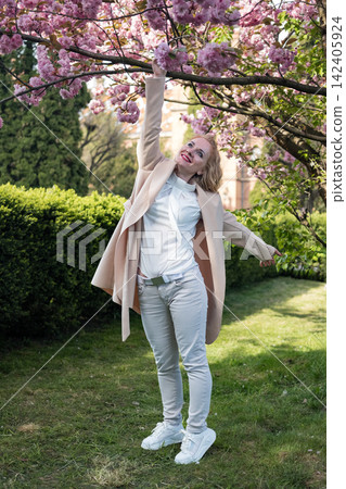 Woman smiling and happily reaching for delicate pink cherry blossoms on a tree branch, celebrating the beauty and joy of springtime outdoors in a park Woman smiling and happily reaching for delicate pink cherry blossoms on a tree branch, celebrating the beauty and joy of springtime outdoors in a park 142405924