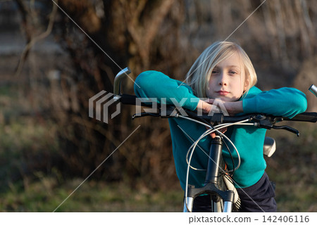 Blonde child leaning on bicycle handlebars, gazing calmly at camera with relaxed, content expression outdoor portrait capturing childhood leisure, innocence and simple freedom Blonde child leaning on bicycle handlebars, gazing calmly at camera with relaxed, content expression outdoor portrait capturing childhood leisure, innocence and simple freedom 142406116
