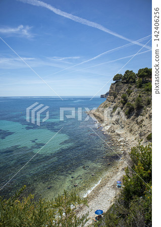 Scenic view of rocky beach cove with clear blue sea, steep cliffs, and green trees under sunny sky Scenic view of rocky beach cove with clear blue sea, steep cliffs, and green trees under sunny sky 142406256