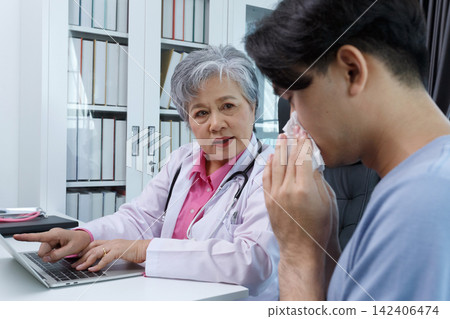 Senior asian female doctor examining and advising young male patient with cough, sore throat, and flu symptoms during medical checkup in clinic office. Hospital treatment and consultation service. Senior asian female doctor examining and advising young male patient with cough, sore throat, and flu symptoms during medical checkup in clinic office. Hospital treatment and consultation service. 142406474