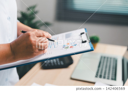 Business professional holding a clipboard and reviewing charts with a pen beside a laptop, representing financial analysis, performance tracking, business planning, strategy, 142406569
