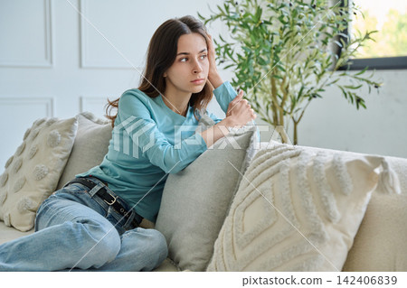 Pensive teen student girl sitting thoughtful on sofa leaning hand Pensive teen student girl sitting thoughtful on sofa leaning hand 142406839