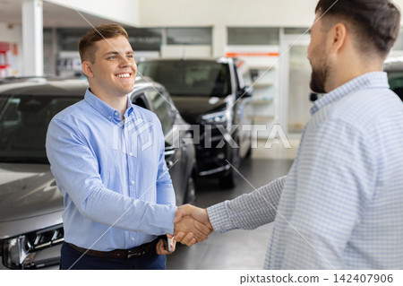 Smiling car sales consultant shaking hands with male customer in dealership showroom 142407906