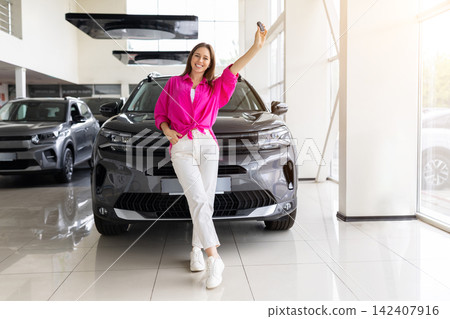 Smiling young woman posing in front of new SUV at dealership showroom, holding key Smiling young woman posing in front of new SUV at dealership showroom, holding key 142407916