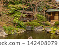 A stone lantern in the sacred pond garden bathed in warm sunlight, and a gazebo standing serenely by the water's edge. March 2024. 142408079
