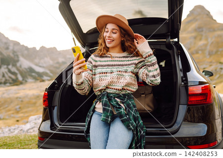 Young traveler woman sits on the rear of her car, smiling and using her phone. Travel and lifestyle Young traveler woman sits on the rear of her car, smiling and using her phone. Travel and lifestyle 142408253