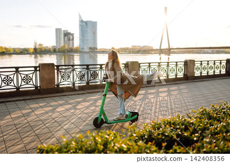 Happy woman driving electric scooter through city. Active life. Ecological transportation concept. Happy woman driving electric scooter through city. Active life. Ecological transportation concept. 142408356