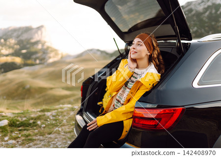 A cheerful traveler enjoying a scenic mountain view while sitting on the trunk. Travelling by car. 142408970