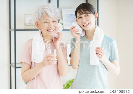 After her exercises, a senior Japanese woman wipes her sweat with a towel and water, while a young Japanese caregiver watches over her. After her exercises, a senior Japanese woman wipes her sweat with a towel and water, while a young Japanese caregiver watches over her. 142408985