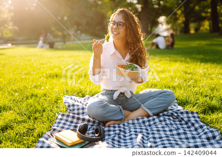 Woman enjoying a poke bowl on sunny day in park while sitting on a picnic by green grass. Vegetarian Woman enjoying a poke bowl on sunny day in park while sitting on a picnic by green grass. Vegetarian 142409044