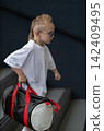 Determined young boy with a stylish mohawk hairstyle and sunglasses walks down modern concrete stairs carrying a black and red sports duffel bag in an urban outdoor setting. 142409495